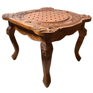 May include: A handcrafted, square wooden table with a carved floral design around the edges and a circular, perforated center. The table has four curved legs and is finished in a warm, natural wood tone.