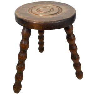 May include: A dark brown wooden stool with three legs. Each leg is made of a series of rounded knobs. The top of the stool has a circular design.