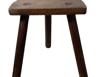 May include: A dark brown wooden stool with three legs. The stool has a rounded top and is made of rough-hewn wood.
