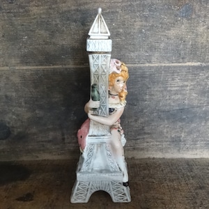 May include: A porcelain figurine of a girl sitting on top of a white Eiffel Tower. The girl is wearing a pink dress and a white hat. The Eiffel Tower is decorated with black lines.