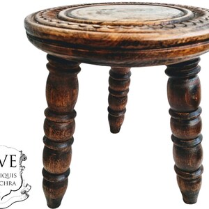 May include: A dark brown wooden stool with a round top and three turned legs. The top of the stool has a carved design.