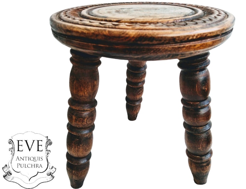 May include: A dark brown wooden stool with a round top and three turned legs. The top of the stool has a carved design.
