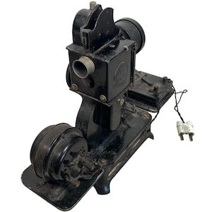 Puede incluir: Un proyector de cine vintage negro con un altavoz redondo y un cable con un enchufe. El proyector tiene una carcasa de metal y una lente en la parte delantera.