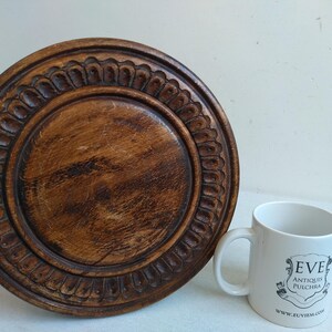 May include: A round wooden pedestal with a carved design around the edge. The pedestal is brown and has a natural wood finish. A white mug with the text "EVE ANTIQUIS PULCHRA WWW.EVEITEM.COM" is in the foreground.