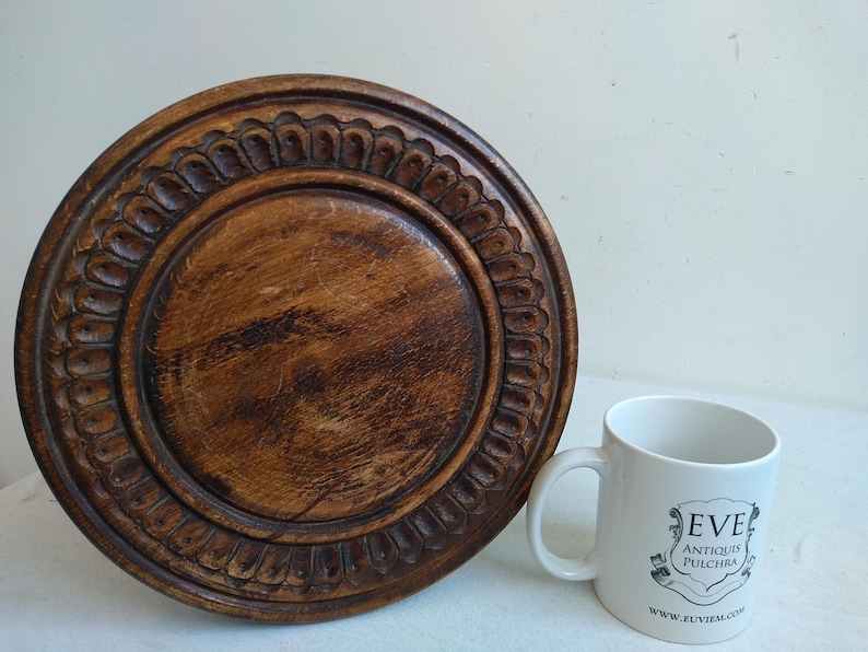 May include: A round wooden pedestal with a carved design around the edge. The pedestal is brown and has a natural wood finish. A white mug with the text "EVE ANTIQUIS PULCHRA WWW.EVEITEM.COM" is in the foreground.