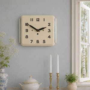 May include: A square, cream-colored wall clock with black numerals and hands. The clock has a blue stripe on each side. The clock is mounted on a light blue wall. The time is approximately 10:10. Home decor.