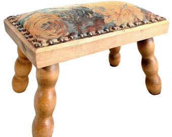 May include: A small wooden footstool with a floral patterned fabric top and turned wooden legs. The fabric is a tapestry with shades of blue, orange, and brown.