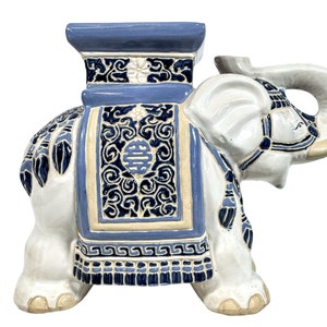 May include: A white ceramic elephant garden stool with blue and gold painted details. The elephant is decorated with a traditional Chinese design and the word "Double Happiness" in Chinese characters. The stool has a square top.