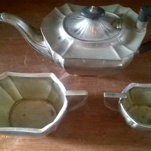 May include: A silver teapot, creamer, and sugar bowl set with a geometric design. The teapot has a black handle.