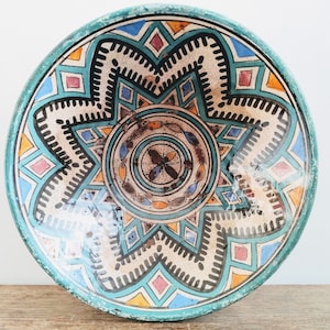 Può includere: Un piatto rotondo in ceramica con sfondo blu turchese e intricati motivi geometrici in nero, bianco, arancione e rosa.