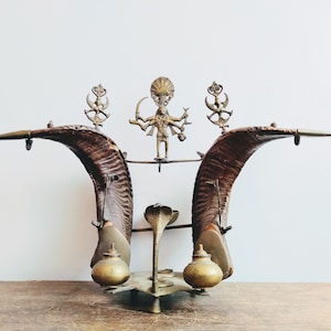 Puede incluir: Un altar de latón con una figura de deidad en el centro, flanqueado por dos cuernos curvos y dos pequeñas ollas de latón a cada lado.