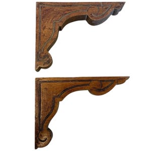 Puede incluir: Dos escuadras de estantería de madera marrón oscuro. Cada escuadra presenta un diseño tallado y ornamentado con bordes curvos y volutas decorativas. Estas escuadras están diseñadas para soportar estantes y añadir un toque rústico a cualquier espacio.