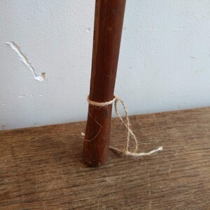 May include: A close-up of a wooden dowel rod with a natural finish. The rod is tied with a piece of twine near the bottom.