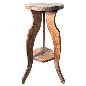 May include: A wooden plant stand with a round top and a lower shelf. The stand has three legs that curve outwards at the bottom.