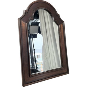 May include: A dark brown wooden framed mirror with an arched top and a bevelled edge. The mirror reflects a white curtain and a glimpse of a room.