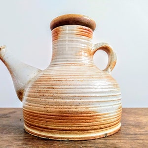 May include: A ceramic pitcher with a spout and handle. The pitcher is primarily white with horizontal brown stripes and a brown rim and stopper. The pitcher is sitting on a wooden surface.