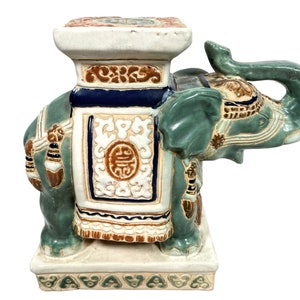 Puede incluir: Un taburete de jardín de cerámica con forma de elefante con un esmalte verde y marrón. El elefante tiene una silla de montar con diseños intrincados y una plataforma elevada en su espalda. La base del taburete está decorada con un patrón floral.