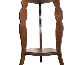 May include: A vintage wooden plant stand with two circular shelves and decorative, curved supports. The stand is made of dark brown wood and features a unique, Art Nouveau design. The top shelf is approximately 12 inches in diameter, and the stand is about 30 inches tall.