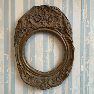 May include: Ornate oval picture frame with a bronze finish. The frame features detailed floral and scrollwork designs around the edges and top. The frame is empty, with a circular opening in the center. The background is a blue and white striped wall.