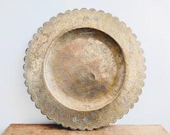 Puede incluir: Una bandeja redonda grande de latón con un diseño intrincado en relieve. La bandeja tiene un borde festoneado y está decorada con un patrón floral.