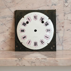 May include: A vintage clock face with a white background, black Roman numerals, and pink accents. The clock face is mounted on a square, dark metal frame. The text "N°9111" is printed on the face.