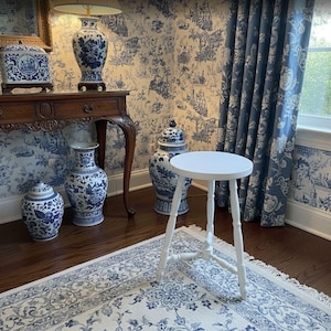 May include: A white, three-legged stool stands on a patterned rug in a room with blue and white decor. The room features blue toile wallpaper, curtains, and several blue and white ceramic vases and jars. A wooden table with a lamp is also visible.