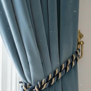 May include: Close-up of a teal blue velvet curtain with a decorative rope tieback. The tieback is a twisted rope design in cream, light blue, and navy blue. A gold-colored metal curtain hook is visible on the right.