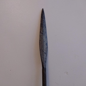 May include: A close-up of a long, narrow, dark gray metal spear point.