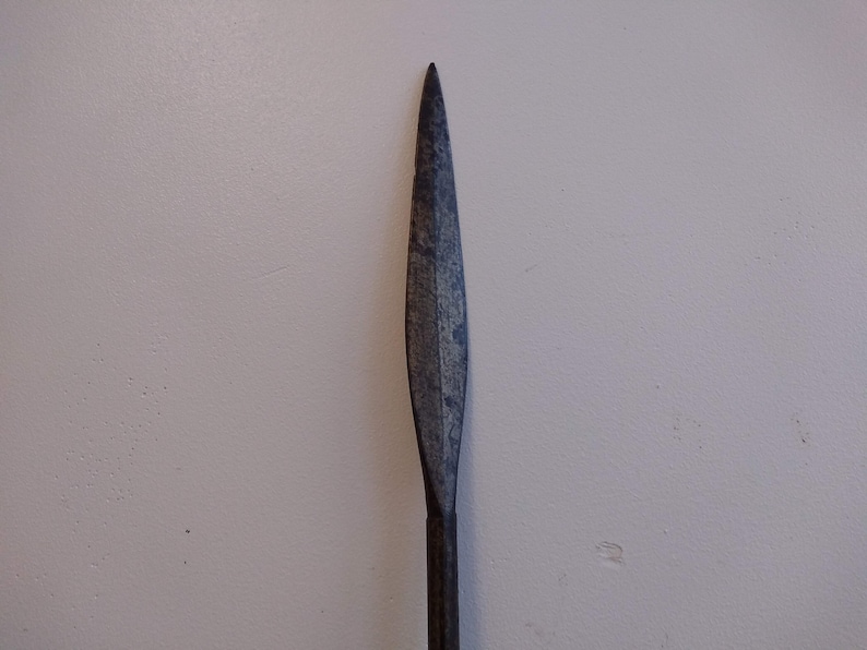 May include: A close-up of a long, narrow, dark gray metal spear point.
