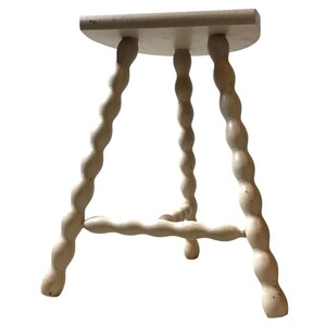 May include: A small, off-white wooden stool with a round seat and three legs. The legs have a unique, twisted design. The stool is painted in a neutral color, suitable for various interior styles.