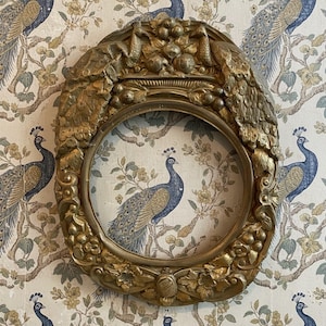 May include: Ornate gold-colored oval picture frame with detailed relief carvings of foliage and fruit. The frame surrounds a peacock-themed fabric. The background features a wallpaper with peacock and floral patterns.