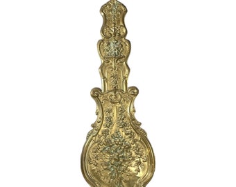 May include: A gold-colored, ornate, decorative pendulum for a clock. The pendulum is shaped like a teardrop with intricate floral designs.