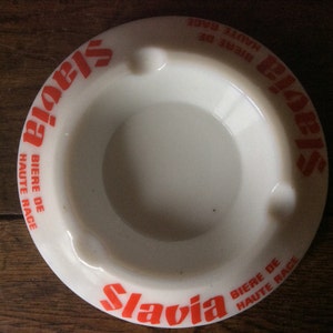 Puede incluir: Cenicero de cerámica blanca con letras naranjas que dicen "Slavia Biere de Haute Race" en ambos lados.