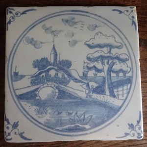 May include: A blue and white ceramic tile with a circular design depicting a landscape scene. The scene includes a bridge, a small building, trees, and a body of water. The tile has a white background with blue details.