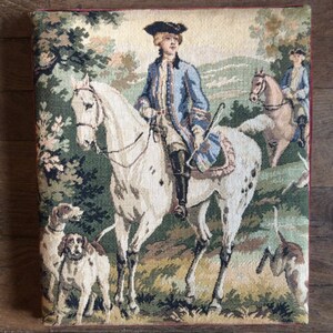 Peut inclure: Une tapisserie représentant un homme à cheval blanc tacheté de brun traversant une forêt. L'homme porte un manteau bleu et un chapeau noir. Deux chiens courent derrière le cheval.