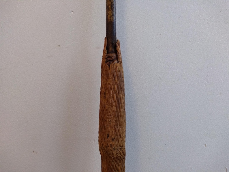 May include: A close-up of the handle of a wooden spear. The handle is wrapped in a woven material and is secured with a metal band.