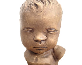 May include: A terracotta bust sculpture of a child with closed eyes and a serious expression. The sculpture is detailed with textured hair and a simple, draped collar.