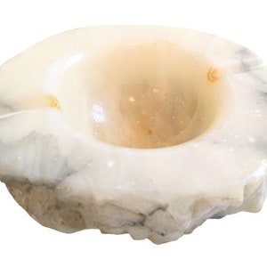 Puede incluir: Un cenicero de alabastro blanco con un borde áspero y natural. El cenicero tiene un cuenco redondo y poco profundo y un color ligeramente blanquecino.