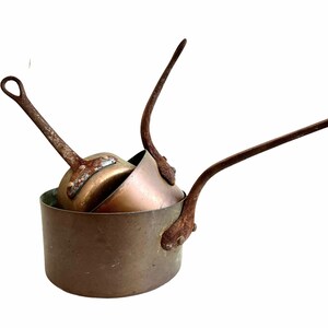 Puede incluir: Tres ollas de cobre vintage con asas largas. Las ollas están anidadas y tienen un aspecto rústico y envejecido.