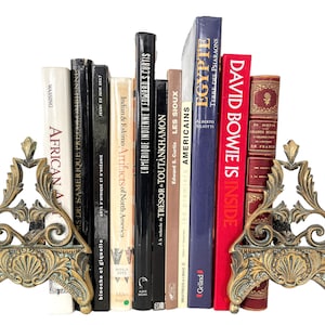 May include: A set of antique brass bookends with ornate floral designs, holding a collection of books with titles like "African Art", "Indian & Eskimo Artifacts of North America", "L'Amerique Indienne d'Edwards Curtis", "Tresor Toutankhamon", "Les Sioux", "Les Premiers Americains", "Egypte", and "David Bowie is Inside".