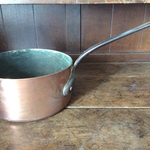 以下が含まれることがあります： 黒い取っ手付きの銅製の鍋。鍋は光沢のある銅の仕上げで、内側に緑色の緑青が付着しています。