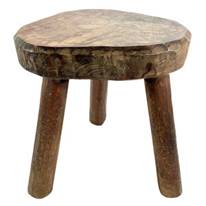 May include: A rustic wooden stool with three legs and a rounded top. The stool is made of dark brown wood and has a natural, weathered finish.