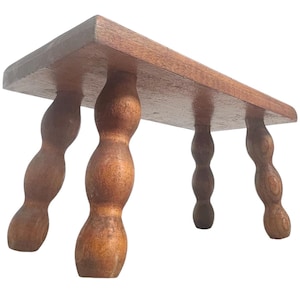 Puede incluir: Un pequeño taburete rectangular de madera con un acabado marrón cálido. El taburete presenta cuatro patas torneadas con un diseño bulboso, proporcionando una base sólida. La superficie superior es lisa y pulida, mostrando la veta natural de la madera. Mide unos 40 cm de largo.
