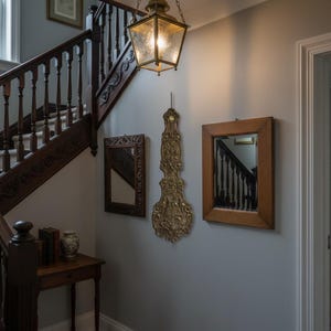 May include: An interior shot of a hallway with a wooden staircase, a hanging lantern, and decorative wall mirrors. A gold-colored, ornate wall clock hangs between two framed mirrors. The staircase has dark wood banisters.