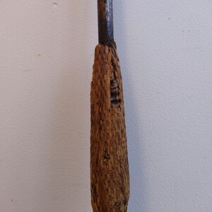 May include: A close-up of a wooden handle for a tool. The handle is made of rough, brown wood and has a worn, rustic look.