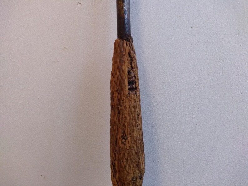 May include: A close-up of a wooden handle for a tool. The handle is made of rough, brown wood and has a worn, rustic look.