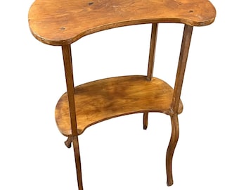 Puede incluir: Una mesa auxiliar de madera de dos niveles, en forma de riñón. La mesa tiene un acabado marrón claro con vetas de madera visibles. El nivel superior es más grande que el inferior. Las patas son delgadas y curvas.