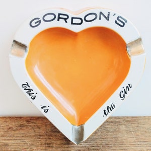 May include: A heart-shaped ceramic ashtray with a white rim and an orange center. The ashtray has the text "Gordon's" in black lettering on the top and "This is the Gin" in black lettering on the bottom.
