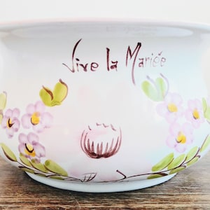 Peut inclure: Pot de fleurs en céramique blanche avec un bord rose et un motif floral. Le pot porte l'inscription "Vive la Mariée" en cursive brune.