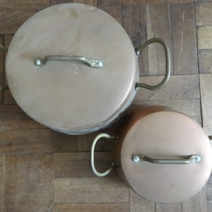 Peut inclure: Deux casseroles en cuivre avec des poignées en laiton. La plus grande casserole est à gauche et la plus petite à droite. Les deux casseroles ont une finition brillante et métallique.